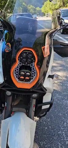 The video shows a close-up, side view of a white KTM Adventure motorcycle parked outdoors. The motorcycle has several stickers, including "KTM", "ADVENTURE", and "ENDURISTAN". It also features a black windshield with stickers, black handguards, a black seat, and a black storage box on the rear. The front of the motorcycle has a prominent headlight assembly with four LED lights. The background shows a blurred outdoor setting, possibly a street or parking area, with hints of foliage and other vehicles. The camera pans slightly to reveal more of the motorcycle's side and front fairing.