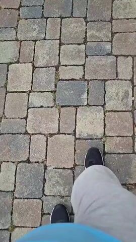 This is a first-person perspective video of someone walking on a stone patio. The camera is pointed downwards at the walker's legs and feet. The person is wearing gray sweatpants and black athletic shoes. The stone patio is made up of rectangular and square pavers in various shades of gray, brown, and red. There are small patches of grass growing between some of the stones. The video shows the person's legs and feet moving as they walk, with their right foot stepping forward at the beginning of the clip and their left foot following.
