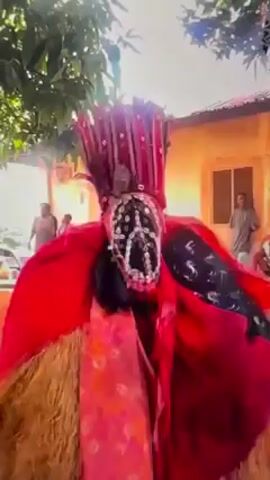 A figure in traditional attire is dancing in a sunny outdoor setting. The costume is predominantly red and gold, with raffia-like material covering the body. The mask features a stylized peace sign made of shells or beads. The figure's movements are energetic and expressive. The background includes trees and a building. The overall impression is that of a cultural performance or festival.
