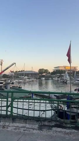 A short video captures a marina scene at dusk. Several boats, ranging from small motorboats to sailboats, are docked in the calm water. A large construction crane is visible in the background, near some buildings and trees. A red flag flies from a flagpole on the right side of the frame. The sky is clear and a soft orange glow suggests the setting sun. In the foreground, a green metal fence runs along the edge of a walkway or dock.