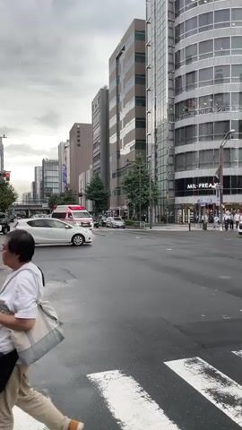 The video shows a street scene in a city with tall buildings. A white ambulance with flashing red lights is driving through an intersection. Several cars are also present on the wet road. The sky is overcast, suggesting it may have recently rained or is about to rain. Pedestrians can be seen on the sidewalk. The overall atmosphere is urban and somewhat busy.