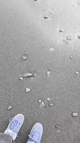The video captures a first-person perspective of someone walking along a dark-sanded beach in Puerto De La Cruz, Spain. The scene is dominated by the dark, wet sand, scattered with pebbles of various sizes. A gentle wave is washing ashore, its white foam creating a stark contrast against the dark sand. The camera angle is fixed on the feet and legs of the individual, who is wearing light blue sneakers with white laces. The individual is walking towards the water, with the waves occasionally reaching their feet. The overall impression is one of a serene and peaceful walk by the sea. The time of day appears to be overcast or early morning/late evening, as the lighting is soft and diffused, and there are no harsh shadows. The weather seems mild, suitable for a beach walk. The location cues, such as the dark sand and the general atmosphere, are consistent with coastal areas in Puerto De La Cruz. There are no visible text elements in the video. The activity is simply walking and experiencing the shoreline. The emotion conveyed is one of calm and connection with nature.