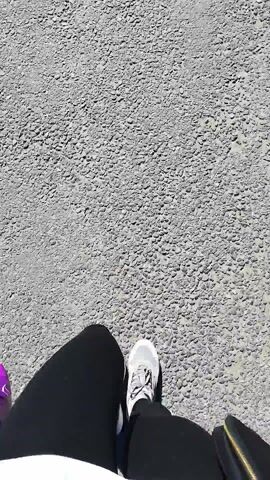 This is a first-person perspective video clip filmed outdoors on a sunny day. The scene is a close-up of a person's feet walking on a rough, gravelly surface, likely a path or road. The person is wearing athletic shoes and dark pants. A shadow is cast on the ground, indicating the time of day is likely late morning or afternoon due to the angle of the sun. No visible text is present. The setting appears to be a casual outdoor environment. The activity is simply walking.