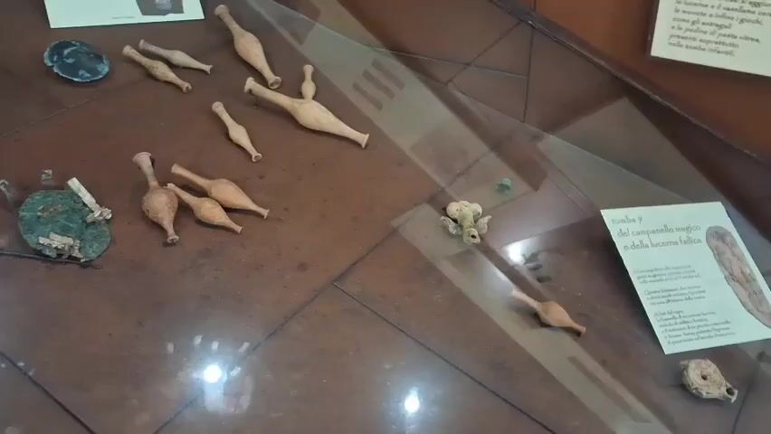 The video shows a museum display case containing ancient artifacts. The objects are primarily small, terracotta vessels, some with long necks and bulbous bases, others with multiple spouts. There are also some metallic objects, one of which appears to be a circular bronze mirror with a green patina, and another small, irregular piece of metal. A larger, lighter-colored stone object is also visible. One section of the display includes a small, light-colored object that resembles a carved figure or fragment, possibly of an animal. A descriptive plaque with Italian text and illustrations is positioned near some of the artifacts. The display case has a glass top, reflecting the lighting and surroundings. The overall impression is of an archaeological exhibition of ancient pottery and other small finds.