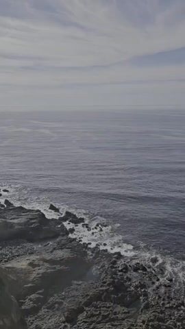 The video shows a high-angle, static shot of a rocky coastline under an overcast sky. The ocean stretches out to the horizon, appearing calm with gentle ripples. Waves break against the dark-colored rocks along the shore. The sky is light gray, with faint streaks of lighter clouds.