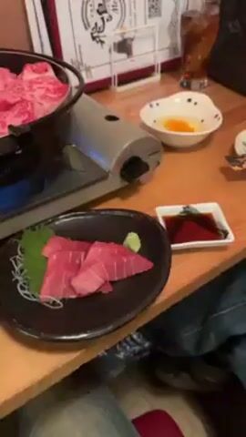 The video shows a table in a restaurant with several dishes. There is a portable gas stove with a black pot on it, which contains sukiyaki with beef, vegetables, and noodles. To the right of the pot is a black plate with slices of tuna sashimi garnished with wasabi and a green leaf, along with a small dish of soy sauce. Next to the pot on the left is a bowl with a raw egg and another bowl of white noodles. The table is made of wood. Some people are visible behind the table.