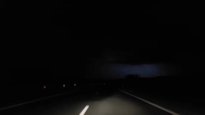The video, filmed at night, shows the perspective of a driver on a dark road. The road is marked by a white solid line on the right, and a white dashed line in the center, which turns into a solid line as the car approaches. Red lights from other vehicles are visible in the distance.