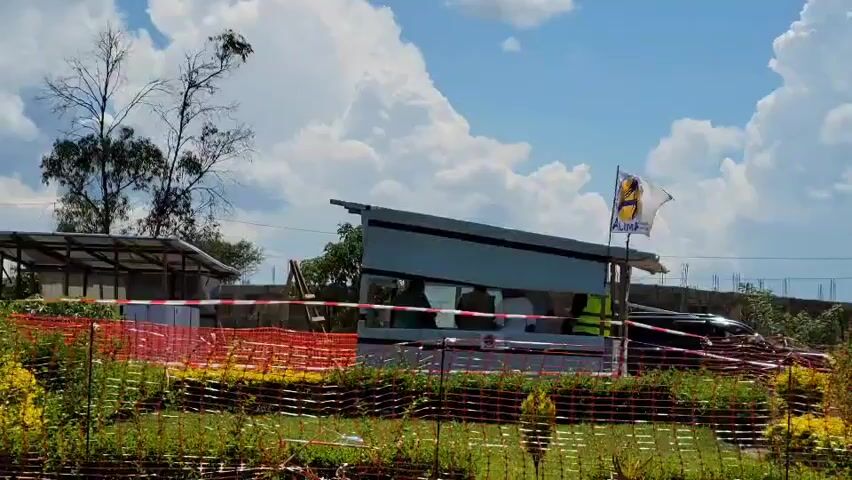 The video captures an outdoor scene under a bright, partly cloudy sky, with large white cumulus clouds against a clear blue. The time of day is sunny daylight.

In the foreground, lush green grass and vegetation are visible, bordered by an orange safety netting or temporary fencing that spans across the frame.

The central focus is a small, light-colored building or shelter featuring an open side, through which several people can be seen inside. Their faces are indistinct due to distance, but at least three individuals are discernible, one of whom appears to be wearing a high-visibility vest. To the right of this structure, a yellow and blue flag with an emblem or text, which is not clearly legible, is flying on a pole. A dark-colored car is partially visible behind the structure and to its right.

In the background, to the left of the main structure, another smaller, possibly roofed shelter or shed is present. Bare trees and distant greenery complete the backdrop. The scene suggests an open area, possibly a site perimeter or a resting point, in Tchere, Democratic Republic of the Congo. The people inside the structure appear to be gathered, but no specific activity or event is clearly evident.