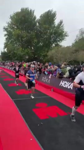 The video shows athletes running a race on a red-carpeted track lined with spectators and banners. One banner features the "Hoka" logo, and another reads "Weymouth Town Council." A person is using their phone to record the athletes crossing the finish line, which displays race times on a digital display. The display lists names such as "Laurence Murphy," "Harry Smith," and "Joseph Musker," along with their times.