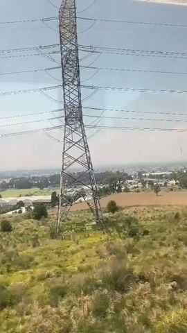 A daytime video from inside a train shows the view from the window as it travels past utility poles. The area outside the train is mostly flat, grassy, and dry. In the distance, a city can be seen with buildings, trees, and fields. The sky is overcast and hazy. The camera focuses on the utility poles and wires as they pass by the train.