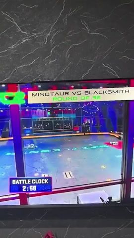 This is a video of a robot combat competition. Two robots, Minotaur and Blacksmith, are in the arena. The battle clock shows 2:57 remaining. The arena is circular with a metallic floor and various obstacles. The crowd is not visible. The video is shot from an elevated angle, looking down at the arena. The lighting is a mix of stage lights and arena lighting. The robots are in motion, with one robot, Minotaur, in the center of the arena and the other, Blacksmith, in the background.