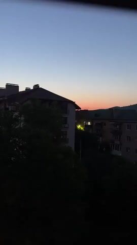 A timelapse video shows a twilight sky over a residential area. The sky is a deep blue, fading to a pale orange at the horizon. Silhouetted buildings and trees fill the foreground, with the lights of a few windows illuminated. Mountains are visible in the distance.