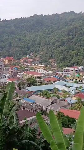 A high-angle drone shot pans across a dense residential area nestled at the foot of a heavily forested hill. Buildings with a variety of colored roofs, predominantly red and blue, are clustered together, with some multi-story structures visible. In the middle ground, a cleared area is set up with white tents and appears to be hosting an event, with small figures, possibly people, gathered on the field. Banana leaves frame the lower portion of the shot, indicating the presence of lush vegetation. The sky is overcast, contributing to a muted lighting effect.