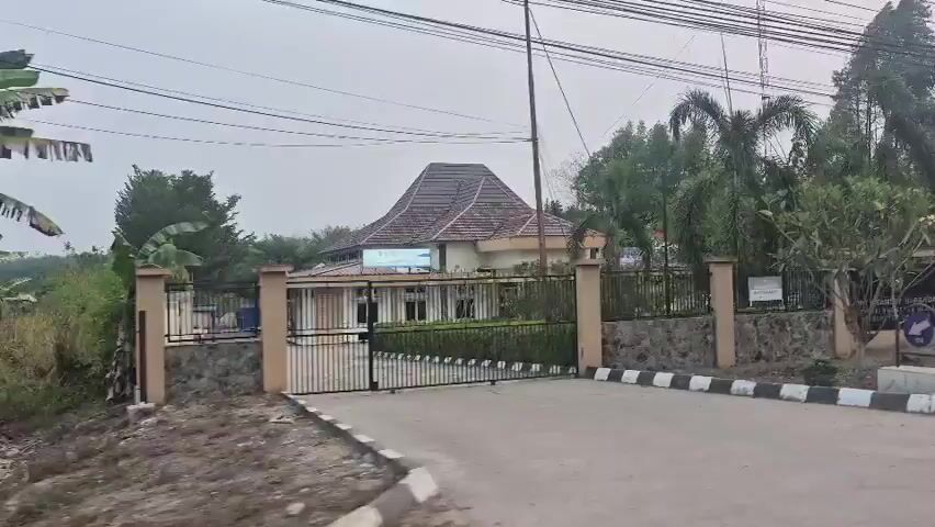The video shows a moving shot of a street with buildings on the right. The buildings appear to be part of a complex or campus, with a fence and gate visible at the entrance. The architecture is of a tropical style with tiled roofs and columns. There are palm trees and other lush vegetation surrounding the buildings. A signpost is visible with text in a foreign language. The sky is overcast. A few cars are parked in front of the buildings. The road in front of the complex is paved and has a black and white striped border.