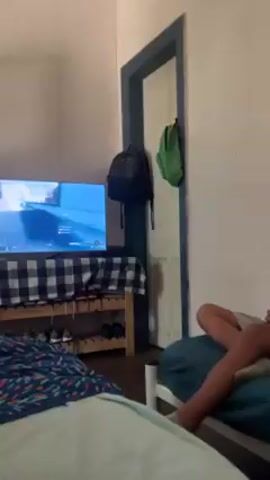 The video shows a boy sitting cross-legged on a bed in a bedroom, playing a video game on a TV. He is wearing glasses, a gray t-shirt, and gray shorts. He holds a blue video game controller. The bed is covered in a blue blanket and a white pillow. Another bed, with a patterned blanket, is visible in the lower left corner of the frame. The TV is on a wooden shelf covered with a black and white checkered cloth. The video game displayed on the TV screen has a blue and white interface. A black backpack and a green bag hang on the wall near the door. The walls are white, and the door has a blue frame. The overall lighting in the room is moderate. The camera moves slightly during the recording.
