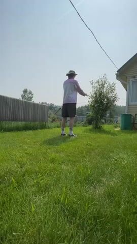 A person wearing a hat, a light-colored jacket with purple accents, dark shorts, white socks with black stripes, and black and white sneakers is performing a flip in a grassy yard. The person is in mid-air, upside down, with their legs bent. They then land on their hands and knees. The background includes a wooden fence, a small tree, a house with yellow siding, a green garbage bin, and a clear blue sky with a wire running across it.