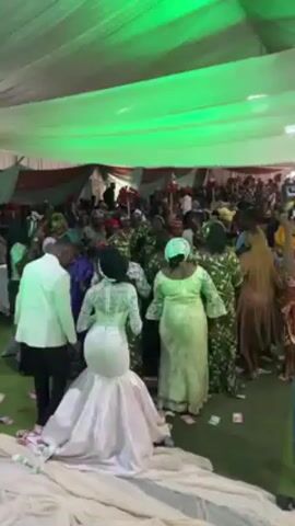 Inside a wedding reception venue, a bride in a white, mermaid-style gown with intricate back detailing is surrounded by guests, many of whom are women in green patterned dresses and head coverings. A man in a white suit is standing beside the bride, holding her hand. There are scattered bills on the green carpet, and the room is decorated with green and red fabric draping from the ceiling. The atmosphere is lively with people moving and talking.