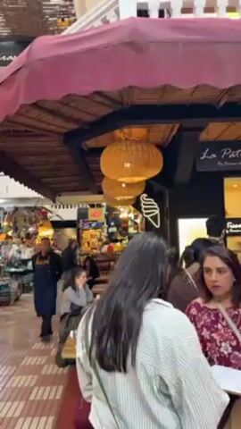 The video shows a busy indoor market. People are walking past various stalls laden with goods, including teapots, lanterns, and other decorative items. Some people are sitting at tables near a restaurant with wicker lamps hanging above. The market has a two-story structure with shops on both levels, and the floor is tiled in a red and white checkered pattern.