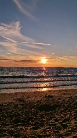 The video shows a sunset over the ocean. The sun is setting in the distance, casting a warm glow over the water and sky. Waves are gently rolling onto the beach, and there is a sandy shore in the foreground. The sky is a mix of blue and orange, with wispy clouds.