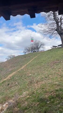 From under the overhang of a structure, a view is captured of a grassy hill dotted with trees. Near the crest of the hill, a Bulgarian flag stands tall, swaying gently in the breeze. Several picnic tables sit under the shade of a bare tree. A narrow dirt path winds its way up the slope. The sky above is a mix of blue and white, with fluffy clouds scattered across it. The landscape has a raw, natural feel, conveying a sense of rural or countryside charm.