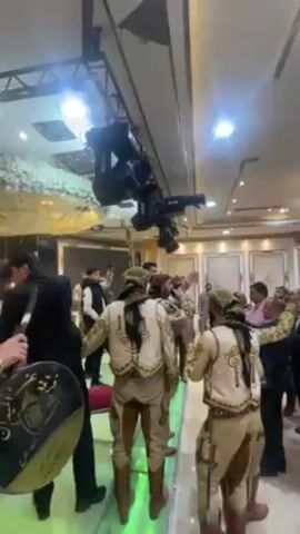 The video shows a gathering in a decorated hall, possibly a wedding or a celebration. A group of performers in traditional clothing, including men wearing ornate vests, baggy pants, boots, and headscarves, stand on a slightly elevated platform. They appear to be part of a musical ensemble, as some are holding instruments, including a large drum with Arabic script. The hall is filled with people, some of whom are clapping and cheering, indicating an enthusiastic response to the performance. A large professional camera rig hangs from the ceiling, suggesting the event is being professionally filmed. The hall is decorated with gold accents and warm lighting.