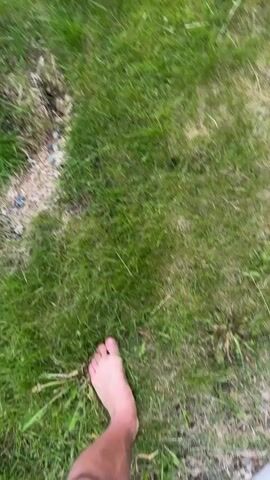 The video shows a first-person perspective of someone walking barefoot on a grassy area. The person's feet are visible as they step on the grass, with the motion blur indicating movement. In the upper left corner of the frame, there is a patch of disturbed ground with some lighter-colored material and small dark objects. The rest of the scene is dominated by green grass, with some weeds or other plants interspersed. The lighting appears to be natural daylight.