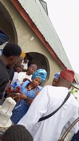 Here's a concise description of the scene depicted in the video:

**Scene:** A group of people are gathered outside a building. The focal point is a woman wearing a blue dress and matching headwrap, being assisted or carried by several individuals.

**People:** There are at least 8 people visible. Several men are wearing traditional Yoruba attire, including caps and white robes. Others are dressed in more modern clothing. The woman in blue appears to be the center of attention.

**Objects:** A white decorated bag is visible. There's also a portion of a white decorated vehicle visible in the background. The building behind the group has a grey roof and arched doorway.

**Actions & Interactions:** People are assisting or possibly carrying the woman in blue. There's an element of support or even celebration surrounding her. Several individuals are looking at her and interacting with her, perhaps to help her.

**Environment:** The setting appears to be outdoors, possibly at an event like a wedding. The building suggests a formal or community setting. The sky is overcast.