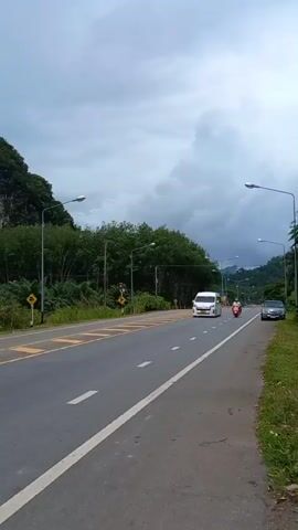 The video shows a road with a white van driving on it. Behind the road are several large, steep rock formations covered with lush green trees and vegetation. The sky is overcast and gray. The road has yellow and white lines, and there are streetlights along the side.