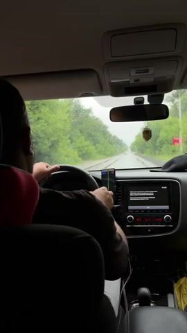The video shows the interior of a car from a rear passenger's perspective. A person wearing a dark shirt and a red headrest is driving. The steering wheel is visible, along with a smartphone mounted on the dashboard displaying navigation. The car's infotainment system is also visible. Outside the car, it is raining and the road is wet, with trees lining the sides. Another car is visible in the distance on the road ahead.