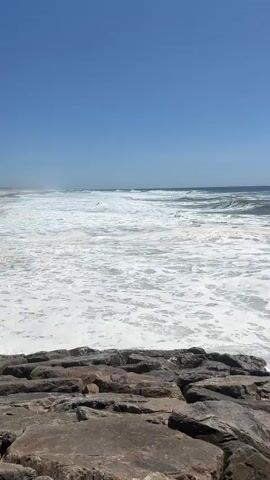 The video shows a view of the ocean from a rocky shoreline. The sky is a clear, bright blue, indicating a sunny day, likely midday given the intensity of the light. The ocean is a vibrant blue-green, with white foam from crashing waves dominating the foreground and middle ground. The waves are moderately sized, rolling in towards the shore and creating a dynamic and active scene.

In the foreground, a collection of large, weathered rocks forms a natural barrier or jetty. These rocks are varied in color, ranging from dark gray to reddish-brown, and are rough in texture, showing signs of erosion from the constant interaction with the sea. They anchor the composition and provide a sense of solidity against the fluid motion of the water.

In the distance, the ocean stretches out to the horizon, where it meets the sky. More waves are visible, appearing smaller and less defined as they are further away. There are no people or man-made structures immediately visible in the frame, emphasizing the natural beauty of the coastal landscape. The overall impression is one of a lively and refreshing day at the beach, with the sound of the waves likely being a prominent feature of the experience. The location is identified as Torreira, Portugal, suggesting a coastal setting typical of the region.