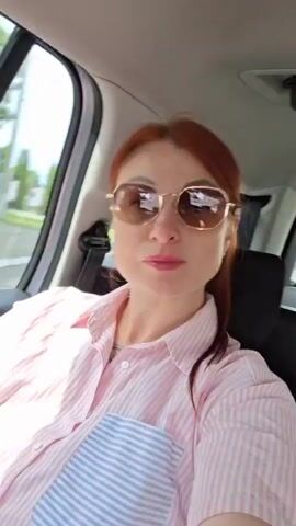 A woman with red hair, wearing a pink and white striped shirt, sunglasses, and pink lipstick, is in the passenger seat of a car. She is making a kissing face at the camera. In the background, out the window, trees and buildings are visible. A white plastic bag with blue writing is on the seat next to her. Another person with curly hair is visible in the back seat. The woman waves to the camera.