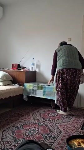 A woman wearing a headscarf, vest, and patterned pants walks toward a door in a bedroom. The room contains a bed, a bedside table, a small table covered with a tablecloth, and a rug. A window with blinds is visible, as is an air conditioner mounted on the wall. A pan with food is visible in the foreground. The video then pans to show the room from a different angle.