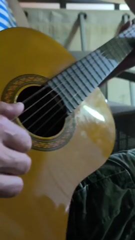 A close-up shot shows a person playing a classical guitar. The guitar is light brown with a decorative rosette around the soundhole. The person's hands are visible, with one hand strumming the strings and the other fretting. The person is wearing a striped shirt and camouflage pants. The background is blurred but appears to be indoors with some furniture and possibly other people.