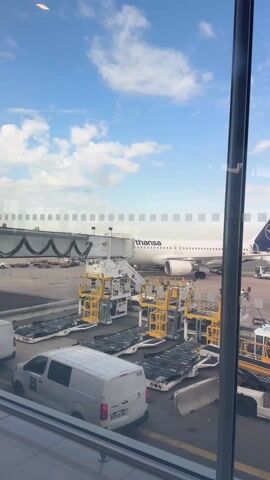 The video displays an active airport tarmac scene under a bright, partly cloudy sky with visible contrails. In the foreground, a white Lufthansa airplane with its distinctive blue crane logo on the tail fin is parked, connected to a jet bridge. The word "Lufthansa" is clearly visible on its fuselage.

Surrounding the aircraft are various airport ground vehicles and equipment. Several yellow and white baggage loaders are positioned, and a white van with "V19" and a "Peugeot" logo, bearing the license plate "BT-052-ZK," is parked nearby. Other ground service equipment, some marked "GEN," and mobile stairs are also visible. Airport ground crew members, identifiable by their orange vests, are moving across the tarmac. A yellow sign marked "X.5" stands on the ground.

The camera pans to the right, revealing more of the vast tarmac and the clear blue sky beyond. The scene captures a typical bustling airport environment, likely during aircraft turnaround operations at what is indicated to be Mauregard, France.