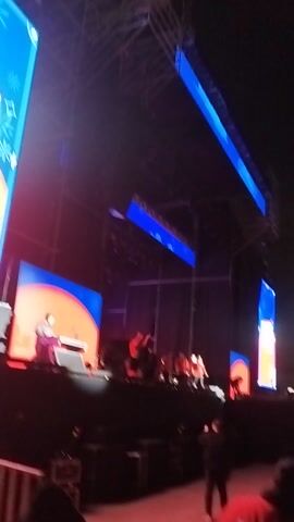 This video captures an indoor event, likely a concert or stage show, at night in Maipú, Chile. The scene features a large stage illuminated by bright lights, with prominent blue and red accents. Several large screens display graphics; one screen on the lower left shows part of a festive design with stars, and possibly the text "THIS SHOW" or similar, while other screens further back show a person on stage. The stage itself has a layered structure with overhead lighting rigs.

A group of people, presumably performers or presenters, are visible on the stage, engaged in an activity that isn't fully clear but seems dynamic. One person on a screen appears to be at a table or desk. The video is filmed from the audience's perspective, looking up towards the stage, capturing the energetic atmosphere of the event.