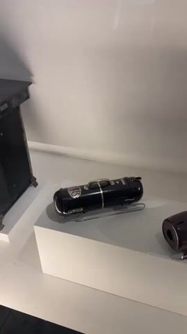 The media file shows a display of vintage vacuum cleaners. The subjects are the vacuum cleaners themselves, which are the primary objects of interest. They are arranged on a white, stepped display platform in what appears to be a museum or exhibition setting. The scene is lit by artificial light, suggesting an indoor environment, possibly a gallery.

There are at least three distinct vacuum cleaners visible. The first is a black, cylindrical model with a chrome handle and accents, accompanied by cleaning attachments. Next to it is a blue and maroon vacuum with a distinctive coiled hose. The third, further to the right and partially obscured by a glass barrier, is a more classic upright vacuum with a metal body, wheels, and a curved handle. The overall atmosphere is one of historical display, showcasing the evolution of household appliances. There are no people visible in the media file, and no weather conditions can be inferred. The location context of Bern, Switzerland, is provided but not visually evident within the frame. No text is visible on the vacuum cleaners or their surroundings. The notable details focus on the design and condition of the vintage appliances.
