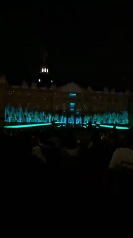 A nighttime outdoor scene shows a building facade illuminated with a light show. The facade is large and appears to be a government building or similar institution, with a dome structure at the top. Projectors are casting geometric patterns of neon blue, pink, and green light onto the building, creating the illusion of a cityscape with tall, abstract buildings. In the foreground, a crowd of people is silhouetted against the light show, their faces and bodies obscured by the darkness. They appear to be standing and watching the display, suggesting an event or performance is taking place. The overall atmosphere is one of spectacle and entertainment.