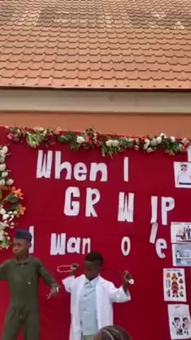 A wide shot shows a group of children standing in front of a red backdrop. The backdrop is decorated with flowers and a sign that reads "When I Grow Up I Want to Be." One child is dressed as a pilot and is standing on the left. Another child is dressed as a doctor and is holding a stethoscope. A third child is standing on the right wearing a white collared shirt. There are pictures of various professions such as firefighter, chef, and business people attached to the backdrop.  The background shows the exterior of a building with a red tile roof.