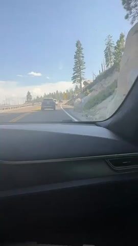 The video shows the view from inside a car driving on a two-lane road. The road is asphalt and has yellow and white lines marking the lanes. There are several cars driving ahead in the same direction. The car is driving on the right side of the road. To the right of the road, there is a steep, rocky hillside with sparse pine trees. The sky is blue with a few white clouds. The sun is shining, casting shadows inside the car. The dashboard of the car is visible in the foreground. There is a yellow sign with a black arrow pointing to the right, indicating a curve in the road. The video is shot from a slightly elevated perspective, looking forward and slightly to the right.