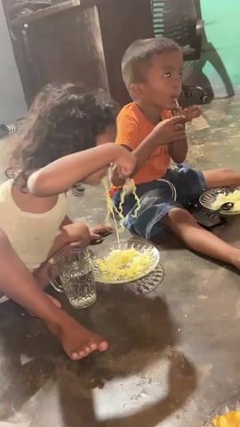 Two young children are sitting on a floor, eating from plates of food. A girl with dark curly hair is on the left, wearing a light-colored top and shorts. She is using a spoon to eat noodles, with some of the noodles hanging out of her mouth. In front of her is a clear glass of water and a plate with noodles. A boy with short dark hair is on the right, wearing an orange t-shirt and denim shorts. He is also eating from a plate of food and looking off to the side. The environment appears to be an indoor space with a concrete floor and furniture in the background. The lighting is dim.
