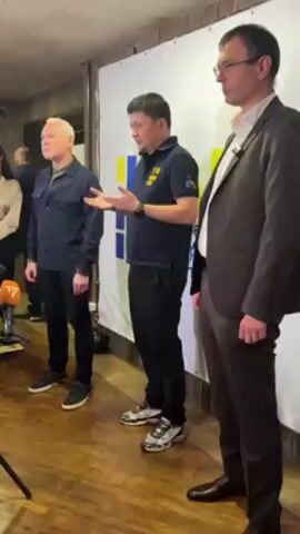 Three men are standing in front of a backdrop that has the colors blue and yellow, which are the colors of the Ukrainian flag. The man in the middle is speaking and gesturing with his hands. He is wearing a navy blue polo shirt and black pants and sneakers. The man on the left has white hair and is wearing a dark blue shirt and black pants and sneakers. The man on the right is wearing a brown suit. There is a microphone to the left of the frame.