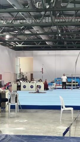 A young female gymnast in a blue and purple leotard performs a floor exercise routine in a gymnasium. She is positioned on a blue mat with judges seated behind her. The gymnast begins in a split position, then transitions to an upright position with her arms raised. She then performs a back handspring, landing back on the mat. The gymnasium features a high ceiling with exposed metal beams and lighting fixtures. Other gymnastic equipment, including parallel bars, is visible in the background. Spectators and coaches are present, with several white chairs scattered around the mat area.