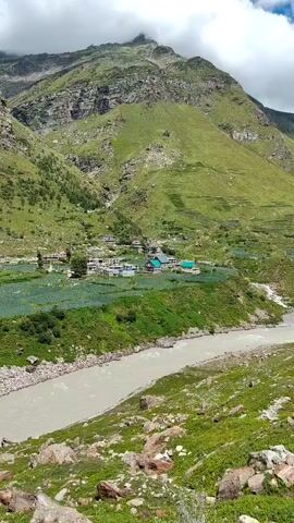 The video shows a sweeping panoramic view of a mountain valley. A wide, muddy river flows through the center of the frame. On either side of the river, steep, green mountains rise dramatically. Patches of vegetation, possibly agricultural fields, are visible on the lower slopes. Small buildings, forming a village, are nestled in the valley. The sky is a vibrant blue with scattered, fluffy white clouds. The camera pans slowly across the landscape, revealing more of the rugged mountain terrain and distant, snow-capped peaks. The overall impression is one of natural beauty and rugged wilderness.