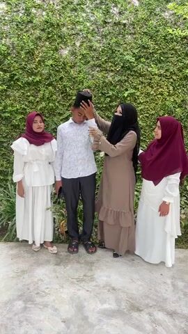 Four individuals stand in front of a lush green wall. From left to right, a young female in a white dress and maroon hijab stands with her hands on her hips. Next to her, a young male in a white patterned shirt and dark pants, wearing a black cap, is touching his head. To his right, a female in a brown abaya and black niqab is adjusting the male's cap. On the far right, another young female in a white dress and maroon hijab stands with her hands clasped. The ground beneath them is a textured concrete surface.