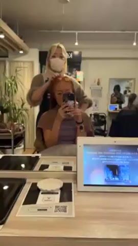 A woman is sitting in a salon chair taking a selfie with her phone while a person stands behind her combing her ginger-colored hair. The person behind her has blonde hair and wears a mask and apron. The salon has other people and salon equipment visible. A small screen in the foreground displays information in Japanese. The woman in the chair is wearing a purple top and has a brown towel around her neck. The person combing her hair uses a pink comb.