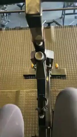 The video shows a first-person perspective of someone using a stair climber in a gym. The stair climber is black and gray, with metal handlebars. The person's legs are visible in gray athletic pants. The camera rises, revealing a view of the gym from the upper level. Below, people are working out on various machines, including treadmills and weight machines. The gym has a gray and black color scheme, with tiled flooring and a high ceiling with exposed beams. The music playing in the background is upbeat.