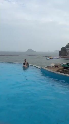 The video opens with a close-up of a swimming pool filled with clear blue water. Two men are visible in the pool, partially submerged and facing away from the camera. The pool edge, a light blue tile border, is prominent in the foreground. The camera then pans right, revealing a wider view of the scene. In the background, a large body of water, likely the ocean, stretches to the horizon under a cloudy sky. A small island or landmass is visible in the distance. To the right of the water, a constructed cliff face with a large waterfall cascading down it dominates the landscape. Above the waterfall, a modern, arched structure with a white roof is visible, and the words "SEA FALLS" are illuminated in large letters. A cartoonish blue sea creature figure is also present near the lettering. Below the waterfall, a sandy beach is visible, with several beach umbrellas and a few people. An excavator is parked on the sand, and construction barriers are present. Further to the right, more land and vegetation are seen, along with a tall communications tower. The video captures a resort or entertainment complex with aquatic features and ongoing construction.