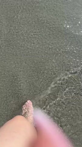 The media file shows a first-person perspective of someone walking in the shallow water at a beach. The sun is glinting off the water's surface, suggesting it is daytime. The sand appears wet and dark, indicating the water is covering it. The person's feet are bare and a light pink polish is visible on their toenails. The water is clear and calm, with small waves gently washing over the person's feet. The overall mood is relaxed and peaceful, evoking the feeling of a day at the beach. No text is visible in the media file.