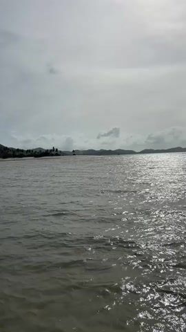 This video shows a view of a body of water, likely the sea, from a slightly elevated perspective, possibly from a boat. The water is choppy, with small waves reflecting the overcast sky. In the distance, a hazy shoreline can be seen with faint outlines of hills or mountains. The sky is filled with clouds, some of which appear brighter, indicating patches of sunlight breaking through. There are no people or distinct objects visible in the foreground or on the water. The overall impression is one of a natural, open environment under a cloudy sky.