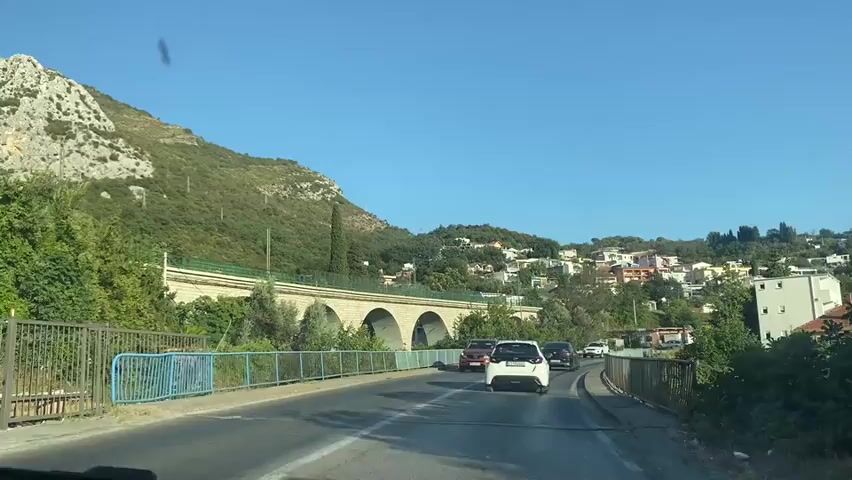 The video shows a forward-facing view from a vehicle driving on a road. On the left side of the road, there is a bridge with multiple arches made of stone. Beyond the bridge, a green, mountainous landscape is visible, with houses scattered on the slopes. The sky is clear and blue. Cars are driving on the road in the same direction as the camera. One car in front is white, and another is black. A dark-colored pickup truck is in the foreground on the left lane. The road has lane markings, and there is a metal railing on the side of the road. Trees and foliage are present on the left and right sides of the road. The time stamp indicates the video is from the early morning.